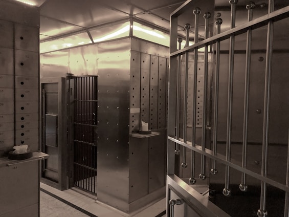 An interior space dominated by metal surfaces and structures, resembling a vault or secure facility. The walls and door are made of metal, with visible rivets and steel bars forming a secure area. There are numbered boxes on the left, possibly indicating storage or safety deposit boxes, and a shelf with a small object or item placed on it.