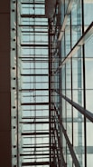The image features a modern architectural structure with a grid-like pattern of glass windows framed by metal beams. The perspective looks upwards, capturing the symmetrical lines and geometric shapes. Soft natural light filters through the translucent panes, casting shadows and creating a serene atmosphere.
