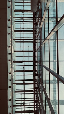 The image features a modern architectural structure with a grid-like pattern of glass windows framed by metal beams. The perspective looks upwards, capturing the symmetrical lines and geometric shapes. Soft natural light filters through the translucent panes, casting shadows and creating a serene atmosphere.