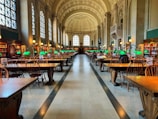 A grand library hall with high arched ceilings and large windows flooding the space with natural light. Rows of wooden tables and chairs are organized neatly, each table adorned with distinctive green desk lamps. Bookshelves line the walls, filled with books, creating an atmosphere of knowledge and study.