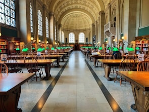 A grand library hall with high arched ceilings and large windows flooding the space with natural light. Rows of wooden tables and chairs are organized neatly, each table adorned with distinctive green desk lamps. Bookshelves line the walls, filled with books, creating an atmosphere of knowledge and study.