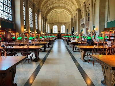 A grand library hall with high arched ceilings and large windows flooding the space with natural light. Rows of wooden tables and chairs are organized neatly, each table adorned with distinctive green desk lamps. Bookshelves line the walls, filled with books, creating an atmosphere of knowledge and study.