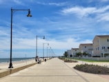 A long, paved boardwalk stretches along a sandy beach with the ocean to the left and a row of street lamps lining the path. On the right, several modern beach houses with balconies are situated, featuring beige and light brown exteriors. The sky is partly cloudy with vibrant blue hues.