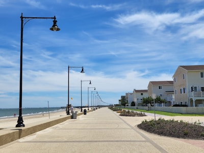 A long, paved boardwalk stretches along a sandy beach with the ocean to the left and a row of street lamps lining the path. On the right, several modern beach houses with balconies are situated, featuring beige and light brown exteriors. The sky is partly cloudy with vibrant blue hues.
