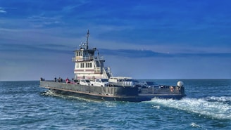 A ferry carrying several vehicles and people travels across a calm sea under a bright blue sky. The water shows gentle waves as the ferry moves, creating a peaceful and serene maritime scene.