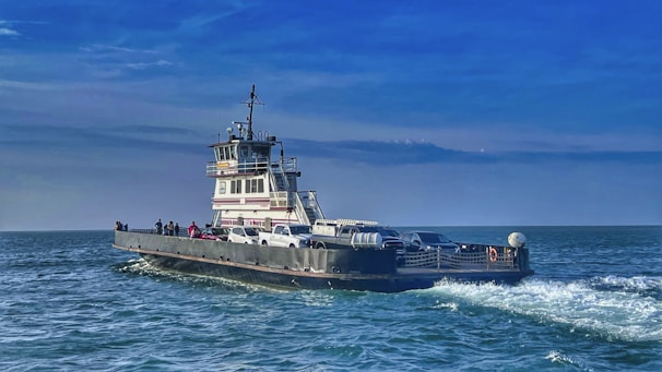 A ferry carrying several vehicles and people travels across a calm sea under a bright blue sky. The water shows gentle waves as the ferry moves, creating a peaceful and serene maritime scene.