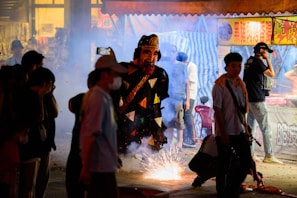 A vibrant street scene during a cultural celebration or festival at night. A large, colorful puppet or figure is central, surrounded by smoke and sparks from fireworks on the ground. People wearing casual clothes and some with masks observe or participate, with a market stall visible in the background.