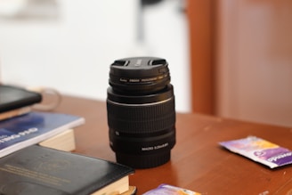 A camera lens is placed upright on a wooden table surrounded by books and some small packets. The lens is black with a silver ring and has clear labeling on its top. The table is slightly cluttered, suggesting a casual setting.