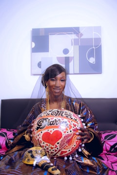 A woman wearing a traditional outfit and veil is holding a decorated item with the words 'Mr. & Mrs. Diarra' and a red heart. The background features a modern geometric painting, and she is sitting on a sofa with pink and black cushions.