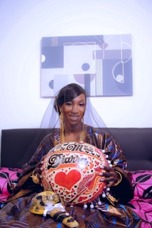 A woman wearing a traditional outfit and veil is holding a decorated item with the words 'Mr. & Mrs. Diarra' and a red heart. The background features a modern geometric painting, and she is sitting on a sofa with pink and black cushions.