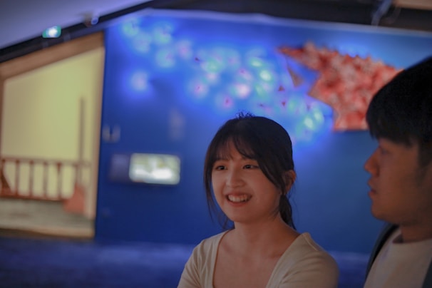 A young woman with dark hair smiles while looking at a young man. They appear to be in a dimly lit room with an abstract, illuminated wall design in the background, displaying soft blue and pink hues.
