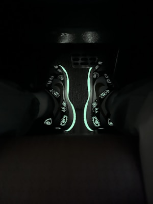 A pair of sneakers with glowing elements are visible in a dimly lit environment. The shoes have distinctive patterns, including eye designs. The glow-in-the-dark features highlight the design against the dark background.