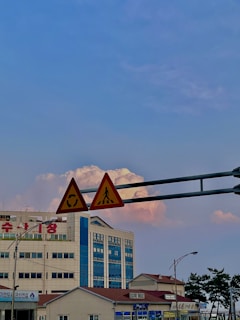Traffic signs are mounted on poles in the foreground, including a pedestrian crossing and a roundabout sign. In the background, there are a modern building with blue and beige sections and red text on it. The sky is partly cloudy with pinkish hues, creating a contrast with the clear blue sky.