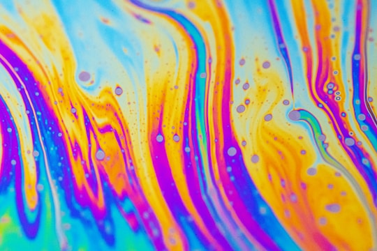 Vivid patterns of swirling colors resembling a psychedelic or abstract art piece, with waves of blue, orange, pink, and yellow blending seamlessly together. Small circular shapes are dispersed throughout, creating an intricate and lively design.