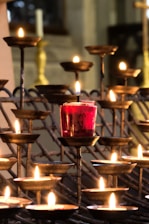A series of metal candle holders with lit candles arranged at varying heights, featuring one prominent red candle in the center. The soft glow of the flames creates a warm and serene atmosphere, with blurred background elements hinting at a church or solemn setting.