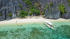 A serene beach with pristine white sand bordered by towering cliffs and lush green vegetation. The clear turquoise water gently laps at the shore. A traditional outrigger boat is anchored nearby, while a few people enjoy the shallow waters and others stroll along the beach.