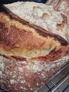 A close-up view of a well-baked artisan loaf of bread with a crusty, golden-brown exterior. The surface is dusted with flour, and the loaf has a prominent scored pattern.