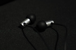 A pair of in-ear headphones with black silicone ear tips and metallic silver casing are placed on a dark surface. The earphones are connected by black cables, and the lighting creates a subtle contrast against the dark background.
