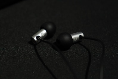 A pair of in-ear headphones with black silicone ear tips and metallic silver casing are placed on a dark surface. The earphones are connected by black cables, and the lighting creates a subtle contrast against the dark background.