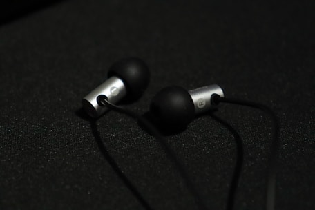 A pair of in-ear headphones with black silicone ear tips and metallic silver casing are placed on a dark surface. The earphones are connected by black cables, and the lighting creates a subtle contrast against the dark background.