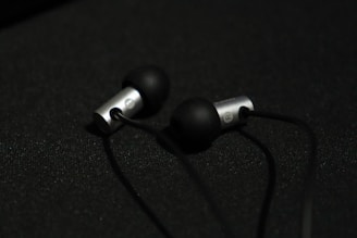 A pair of in-ear headphones with black silicone ear tips and metallic silver casing are placed on a dark surface. The earphones are connected by black cables, and the lighting creates a subtle contrast against the dark background.