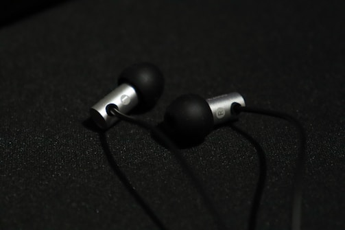 A pair of in-ear headphones with black silicone ear tips and metallic silver casing are placed on a dark surface. The earphones are connected by black cables, and the lighting creates a subtle contrast against the dark background.