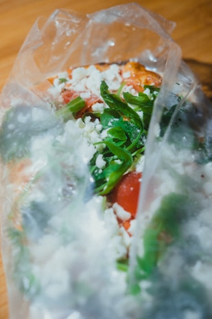 A plastic bag containing fresh ingredients such as spinach leaves, sliced tomatoes, and crumbled feta cheese. The contents appear colorful and appetizing, with the greens and reds contrasting against the white cheese.