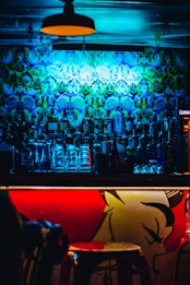 A dimly lit bar with vibrant psychedelic wall art illuminated by blue and green lights. There is a well-stocked bar with a variety of liquor bottles and stacked glassware. The front of the bar counter features bold red and yellow artwork, adding to the vibrant atmosphere.