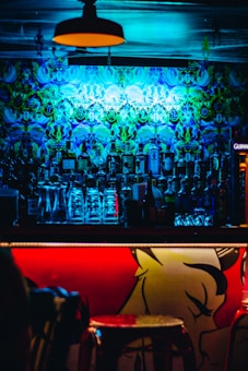 A dimly lit bar with vibrant psychedelic wall art illuminated by blue and green lights. There is a well-stocked bar with a variety of liquor bottles and stacked glassware. The front of the bar counter features bold red and yellow artwork, adding to the vibrant atmosphere.