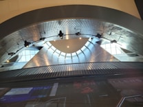 An interior architectural feature with metallic, geometric shapes forming an archway. Spotlights hang from the ceiling, illuminating the futuristic and modern design. Light reflects off various surfaces, creating a dynamic and vibrant atmosphere. The lower part of the image contains text partially visible.