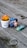 A wooden surface serves as a backdrop for a small arrangement of playful objects. Two cute figurines, a boy and a girl, are positioned next to each other as if kissing. Nearby is a small toy car, colored turquoise with a yellow stripe, and the number 22 on its side. A white pot with the phrase 'Aloe You Vera Much' holds an artificial orange and yellow plant.