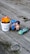 A wooden surface serves as a backdrop for a small arrangement of playful objects. Two cute figurines, a boy and a girl, are positioned next to each other as if kissing. Nearby is a small toy car, colored turquoise with a yellow stripe, and the number 22 on its side. A white pot with the phrase 'Aloe You Vera Much' holds an artificial orange and yellow plant.