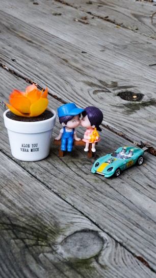 A wooden surface serves as a backdrop for a small arrangement of playful objects. Two cute figurines, a boy and a girl, are positioned next to each other as if kissing. Nearby is a small toy car, colored turquoise with a yellow stripe, and the number 22 on its side. A white pot with the phrase 'Aloe You Vera Much' holds an artificial orange and yellow plant.