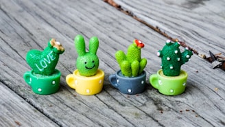 Four small, brightly colored cactus-shaped figurines are placed in miniature teacups. The figurines include a heart with the word 'LOVE,' a bunny face, and two cacti with flowers and polka dots, arranged on a wooden surface.