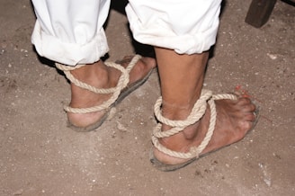 A pair of feet is bound by rope in simple sandals on a dirt ground. The ankles are tied securely with coarse, light-colored rope, and the person is wearing white rolled-up pants.
