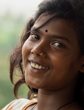 A young woman with dark hair smiles warmly, adorned with a small bindi on her forehead and a nose ring. She has an engaging expression, and her hair flows naturally around her face. The background is a soft blur, suggesting lush greenery.