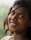 A young woman with dark hair smiles warmly, adorned with a small bindi on her forehead and a nose ring. She has an engaging expression, and her hair flows naturally around her face. The background is a soft blur, suggesting lush greenery.