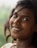 A young woman with dark hair smiles warmly, adorned with a small bindi on her forehead and a nose ring. She has an engaging expression, and her hair flows naturally around her face. The background is a soft blur, suggesting lush greenery.