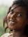 A young woman with dark hair smiles warmly, adorned with a small bindi on her forehead and a nose ring. She has an engaging expression, and her hair flows naturally around her face. The background is a soft blur, suggesting lush greenery.