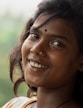 A young woman with dark hair smiles warmly, adorned with a small bindi on her forehead and a nose ring. She has an engaging expression, and her hair flows naturally around her face. The background is a soft blur, suggesting lush greenery.