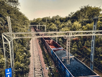 A railway track runs through a lush green forest area, with a long freight train carrying coal traveling on the track. Electrical poles and wires are visible above the train, indicating it is an electrified railway line. The scene is calm, surrounded by dense vegetation.