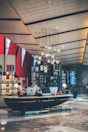 A modern hotel lobby features a sleek, curved reception desk with computer terminals and decorative elements. Above hangs an artistic chandelier with spherical lights. The space has a contemporary architectural design, incorporating geometric shapes and a high ceiling with organic, flowing lines. On one side, shelves display various items, adding a touch of elegance.
