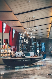 A modern hotel lobby features a sleek, curved reception desk with computer terminals and decorative elements. Above hangs an artistic chandelier with spherical lights. The space has a contemporary architectural design, incorporating geometric shapes and a high ceiling with organic, flowing lines. On one side, shelves display various items, adding a touch of elegance.