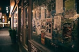 A dimly lit street scene with a storefront lined with various flyers and posters. The posters include advertisements and event announcements, some featuring graphics and bold text. The street beyond is lined with warm lights and a few small potted plants. The overall atmosphere is urban and slightly nostalgic.