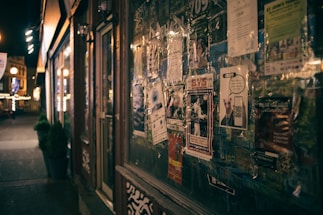 A dimly lit street scene with a storefront lined with various flyers and posters. The posters include advertisements and event announcements, some featuring graphics and bold text. The street beyond is lined with warm lights and a few small potted plants. The overall atmosphere is urban and slightly nostalgic.