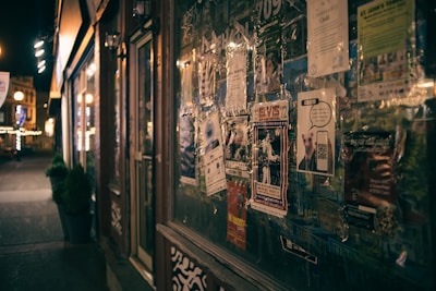 A dimly lit street scene with a storefront lined with various flyers and posters. The posters include advertisements and event announcements, some featuring graphics and bold text. The street beyond is lined with warm lights and a few small potted plants. The overall atmosphere is urban and slightly nostalgic.