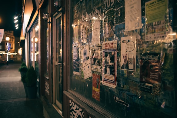 A dimly lit street scene with a storefront lined with various flyers and posters. The posters include advertisements and event announcements, some featuring graphics and bold text. The street beyond is lined with warm lights and a few small potted plants. The overall atmosphere is urban and slightly nostalgic.