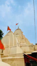 The image features a traditional Indian temple with intricately carved spires and domes, adorned with orange flags. The temple is painted in a light cream color against a clear blue sky.