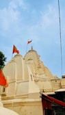 The image features a traditional Indian temple with intricately carved spires and domes, adorned with orange flags. The temple is painted in a light cream color against a clear blue sky.