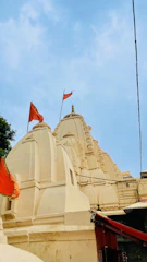 The image features a traditional Indian temple with intricately carved spires and domes, adorned with orange flags. The temple is painted in a light cream color against a clear blue sky.